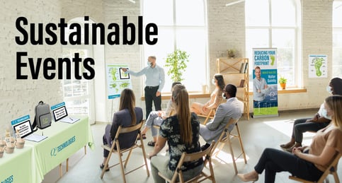 Sunny meeting room where 7 individuals are seated listening to a man give a presentation.  Around the room are various event graphics - a retractable banner, an easel sign, and a table with a backpack and coffee cups on it